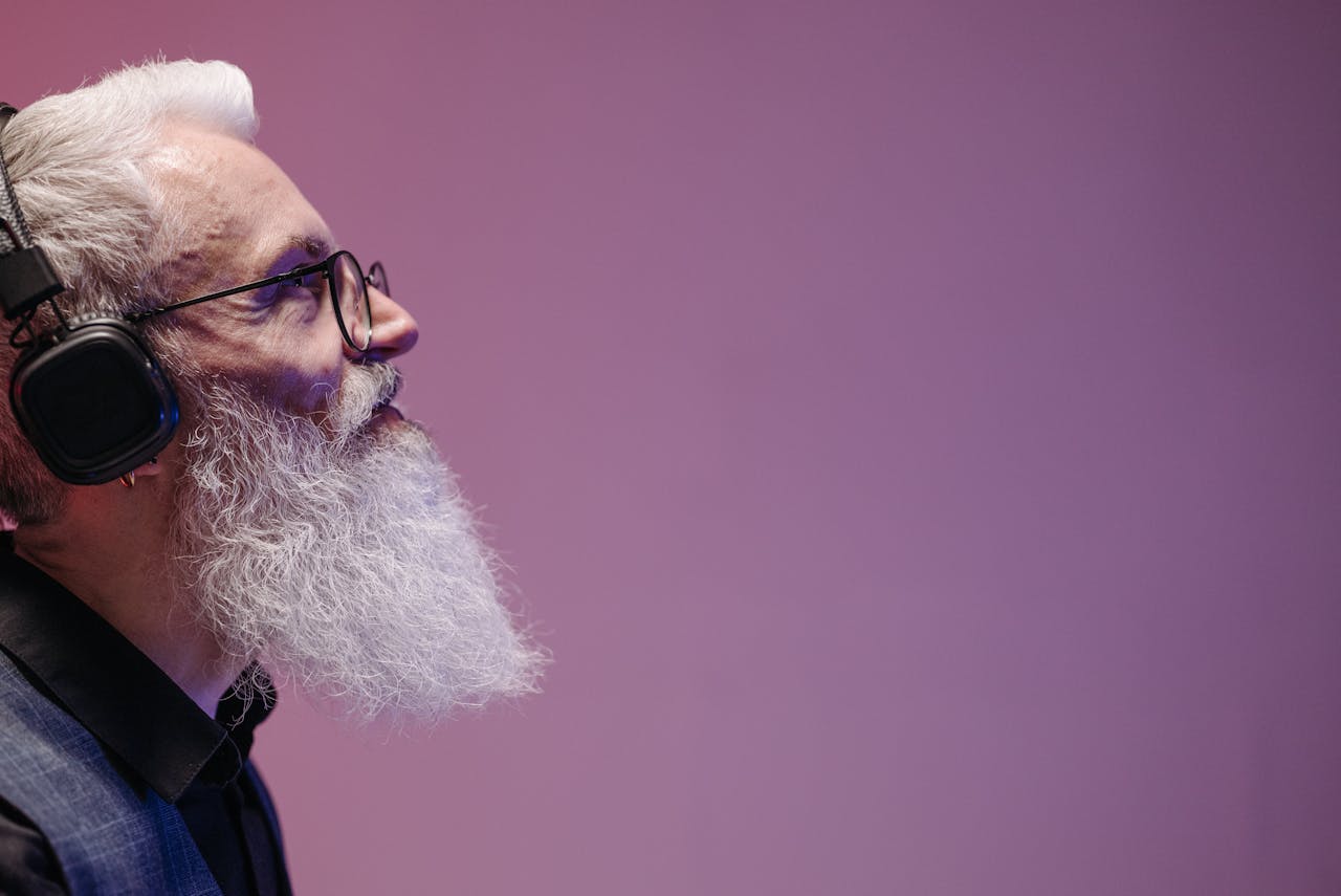Profile of a senior adult man with a long white beard wearing headphones on a purple background.